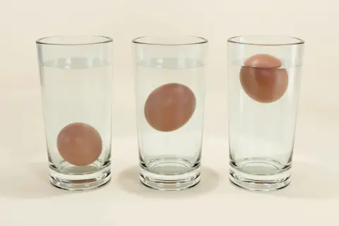 Trois verres d'eau montrent des œufs : un au fond, un flottant au milieu, et un autre flottant à la surface, suggérant des œufs périmés 15 jours.