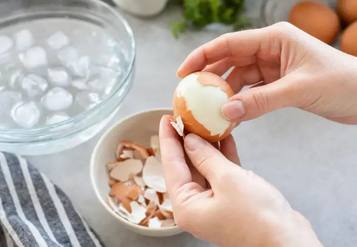 Mains épluchant un œuf dur, prêt pour la cuisson des œufs. Une écumoire d'eau glacée et une coupelle de coquilles sont à proximité.