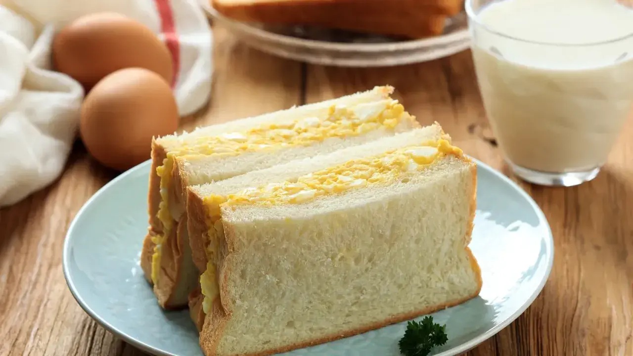 Un délicieux sandwich aux œufs, garni de pain blanc moelleux, servi sur une assiette bleue avec un verre de lait.