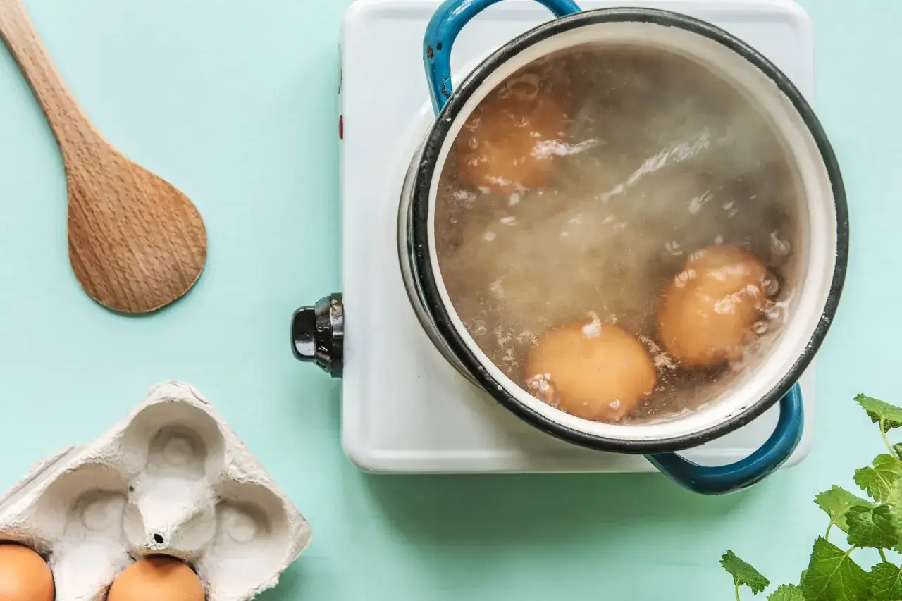 Œufs en cuisson dure dans une casserole bouillante. Une cuillère en bois et une boîte à œufs sont à proximité.